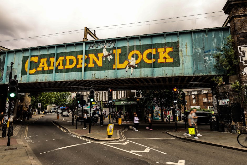 A vintage railway bridge painted in light blue with large yellow lettering spelling 'CAMDEN LOCK' and illustrations of children hanging from the letters. Beneath the bridge, a busy urban street scene is visible with pedestrians on the pavement, some walking and others carrying packages or bags. There are several black and white traffic lights, some illuminated, and a few vehicles, including a white van and cars, driving through the intersection. The scene features shops and buildings with graffiti and street art on the walls, as well as trees and street furniture. This setting is typical for a location involving home relocation or furniture transport, with [COMPANY_NAME]'s removals service closely related to the logistics of loading or unloading items near the Camden area. The overall environment appears lively and active, with a mix of pedestrians, vehicles, and urban infrastructure.
