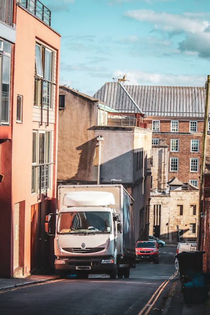 A white box truck from Man with Van Somers Town is parked on a narrow residential street, positioned partly on the pavement and facing uphill. The truck is surrounded by multi-storey brick and stucco buildings with large windows, typical of urban housing. A black car is parked behind the truck, and additional vehicles are visible further down the street. The area appears quiet with clear skies overhead, and the street features yellow road markings. The scene captures the loading process involved in house removals, with the truck ready for furniture transport and packing materials, such as cardboard boxes and blankets, possibly inside it or being prepared for loading. The buildings include modern and older architectural styles, and the environment suggests an active, well-maintained neighbourhood suitable for home relocation services provided by [COMPANY_NAME], supporting efficient furniture transport and moving logistics.