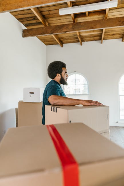 A man with dark curly hair and a beard, wearing a blue T-shirt, is standing inside a room with white walls and a wooden ceiling with exposed beams. He is placing his hands on a large, closed cardboard box that is sealed with red packing tape. Several other cardboard boxes of varying sizes are stacked around him, some on the floor and one directly behind him. The room has two arched windows allowing natural light to illuminate the space, which appears to be in the process of a home relocation or furniture transport. The setting suggests packing and moving activities typical of professional removals, with [COMPANY_NAME], Man with Van Somers Town, specializing in house removals and logistics. The overall scene emphasizes a careful loading process during a move, with attention to packaging materials and spatial organization, supporting seamless furniture transport and packing operations.
