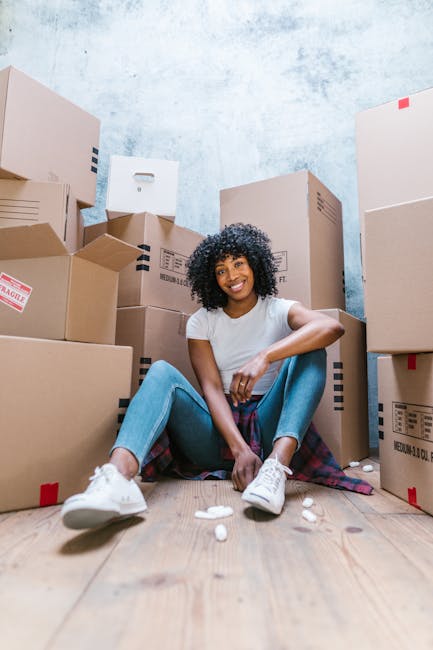 A young woman with curly black hair and a bright smile is sitting on a wooden floor in an inside space surrounded by numerous cardboard boxes of various sizes, some labeled with red and black tape, indicating they are packed for a house move. She is dressed casually in a white T-shirt and blue jeans, with white sneakers, and has a plaid shirt tied around her waist. The boxes are stacked and organized in the background, with some positioned against a textured blue wall. The scene appears to represent the packing stage of a home relocation. The woman’s relaxed pose and the presence of packed boxes reflect a moving and furniture transport process, which aligns with the services offered by Man with Van Somers Town, a professional removals company specializing in house removals and moving logistics in the Somers Town area.