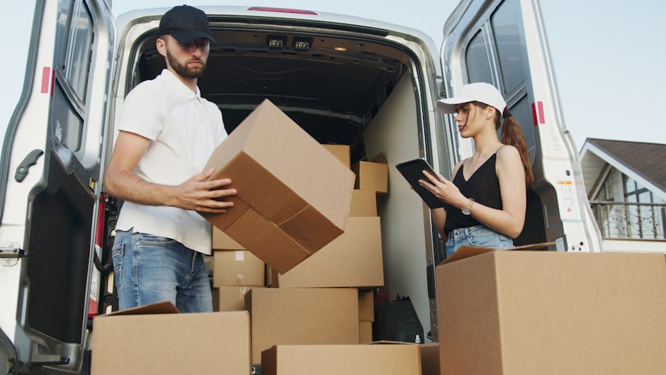 A man and a woman are engaged in a home relocation process outside a residential property, standing next to a large white moving van with its rear doors open. The man, dressed in a white t-shirt, blue