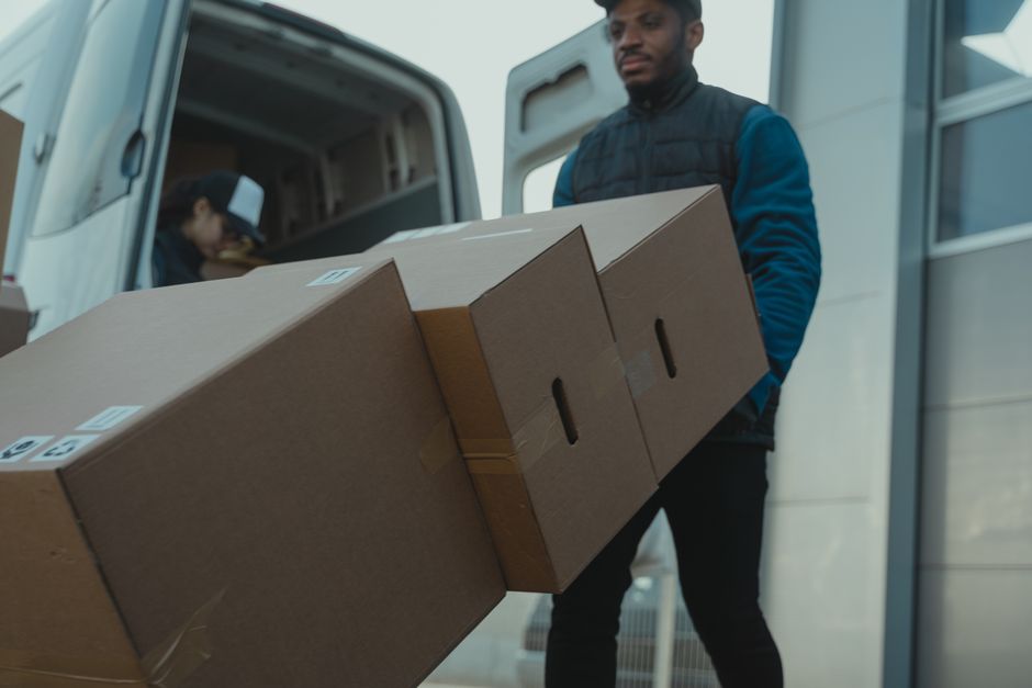 A man and a woman are engaged in a home relocation process outside a residential property, standing next to a large white moving van with its rear doors open. The man, dressed in a white t-shirt, blue