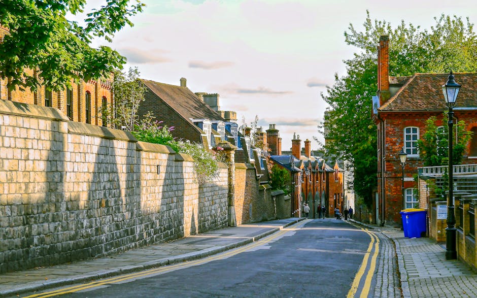 A quiet residential street in Somers Town with a gently curving asphalt road lined by a stone wall on the left and a row of brick houses on the right. The stone wall, constructed from large sandstone blocks, shows signs of weathering, with some greenery and flowering plants visible behind it. The brick houses on the right feature traditional white-framed windows and pitched roofs with chimneys, with some chimneys enclosed in metal caps. A vintage-style street lamp is positioned near the sidewalk, which is paved with concrete slabs. A blue recycling bin is placed near the curb, close to the house entrance. The scene is illuminated by natural, soft daylight, casting shadows on the pavement and facades, with a few pedestrians visible in the distance, suggesting a peaceful day for home relocation and furniture transport activities. This setting reflects a typical environment for moving services by companies such as Man with Van Somers Town, with ample space for loading and unloading household belongings during a home removal or moving process.