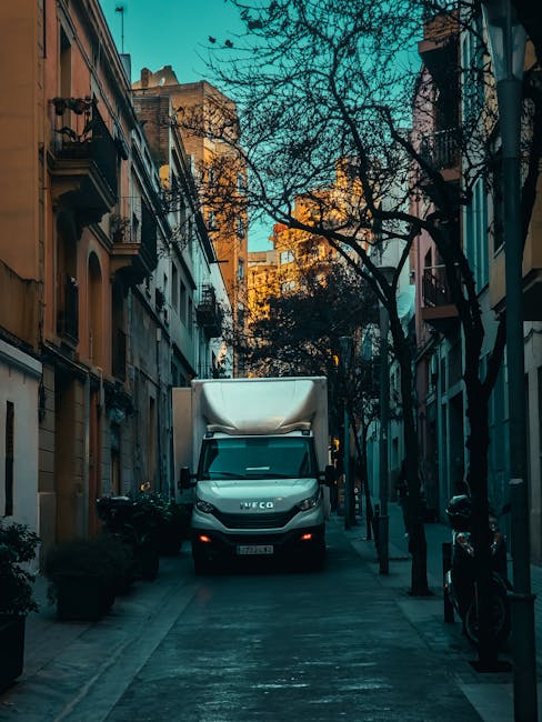 A white Iveco moving van parked on a narrow residential street, with its cargo area covered by a white tarpaulin. The van is positioned near the curb, with the front facing towards the viewer, ready for a home relocation or furniture transport. The street is lined with multi-storey buildings featuring balconies and classic architectural details, with leafless trees lining the pavement. Several cardboard boxes, wrapped furniture, and packing materials are visible inside the van, indicating an active loading process orchestrated by Man with Van Somers Town. A trolley and straps are seen near the van, used to facilitate the furniture moving and loading tasks in the process of packing and moving items into the vehicle for transportation. The lighting suggests early morning or late afternoon, with a clear sky overhead, emphasizing the urban environment where professional removals are taking place.