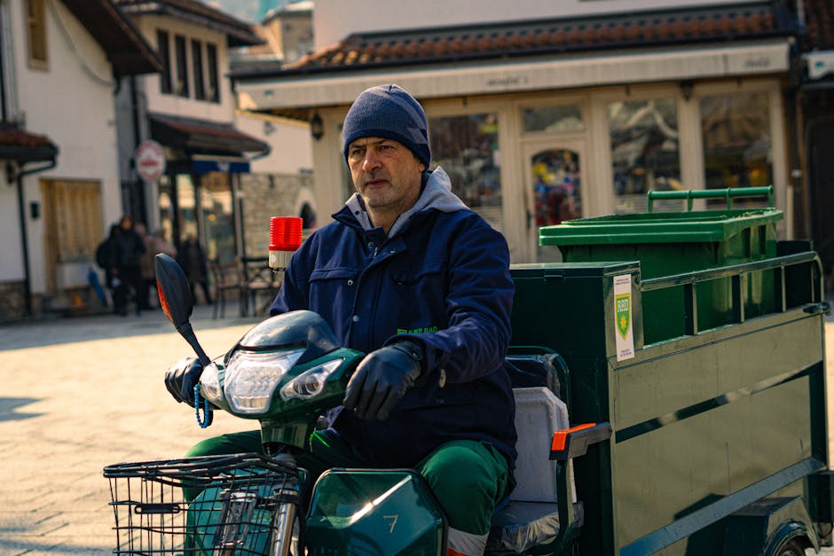 A man wearing a navy blue jacket, grey hoodie, and dark blue beanie hat is seated on a small electric delivery tricycle in an outdoor urban setting. He is holding the handlebars of the vehicle, which is loaded with large green plastic bins and cardboard boxes. The side of the vehicle features a company logo for Man with Van Somers Town, indicating it is used for house removals and furniture transport services. In the background, there are pedestrians, traditional buildings with timber frames and bay windows, and a paved street. The scene suggests a home relocation or moving process, with the man preparing to transport bulky waste and packing materials, possibly during a move or clearance for a property. The lighting is natural, with clear daylight highlighting the scene and the various objects associated with packing and moving.