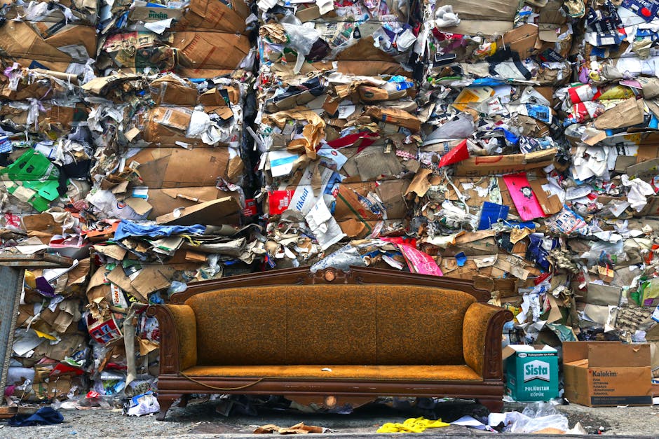 A brown upholstered sofa with wooden framing and carved details is positioned in front of a large pile of assorted waste materials, including crumpled cardboard boxes, paper packaging, plastic wrappers, and damaged packaging materials. The sofa is placed on a sidewalk against a backdrop of a high wall of compressed garbage bags and loose waste, indicating a waste removal or clearance process during a home relocation or furniture transport. Nearby, there are cardboard boxes, some branded, and other debris, with the area illuminated by natural daylight, suggesting an outdoor setting on a street or driveway. This scene illustrates the challenges of managing bulky waste during a house removal, with the presence of a removal company's equipment, such as trolleys or straps, possibly being used to facilitate the loading process. Man with Van Somers Town may provide services to help dispose of such waste efficiently during furniture transport and home relocation.