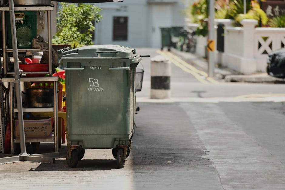 A man wearing a navy blue jacket, grey hoodie, and dark blue beanie hat is seated on a small electric delivery tricycle in an outdoor urban setting. He is holding the handlebars of the vehicle, which is loaded with large green plastic bins and cardboard boxes. The side of the vehicle features a company logo for Man with Van Somers Town, indicating it is used for house removals and furniture transport services. In the background, there are pedestrians, traditional buildings with timber frames and bay windows, and a paved street. The scene suggests a home relocation or moving process, with the man preparing to transport bulky waste and packing materials, possibly during a move or clearance for a property. The lighting is natural, with clear daylight highlighting the scene and the various objects associated with packing and moving.