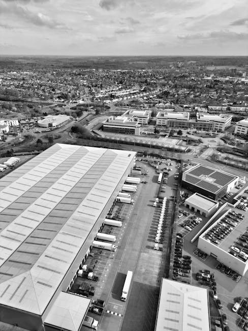 A large aerial view in black and white showing an industrial and commercial area with multiple warehouse buildings and large flat roofs, some with loading bays and trucks parked nearby. The scene includes a parking lot filled with numerous cars, a section with vehicle lanes, and surrounding roads. Beyond the industrial complex, there are smaller buildings, open spaces, and trees extending into the distance, with a cityscape and open landscape visible on the horizon under a cloudy sky. This image of a busy logistics environment relates to house removals and furniture transport, highlighting the logistics involved in home relocation and packing logistics, as presented by Man with Van Somers Town.