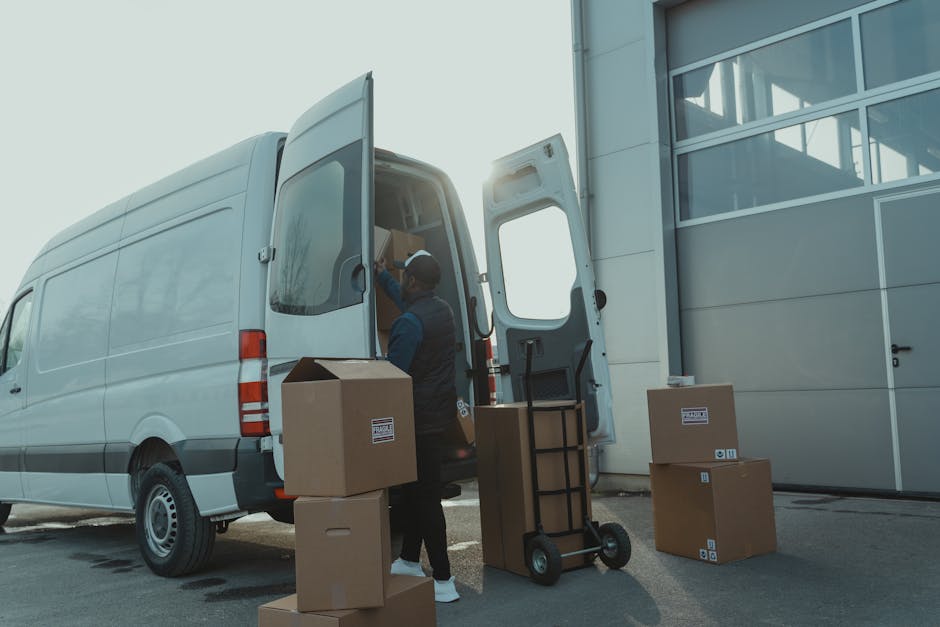 A professional mover from Man with Van Somers Town is loading cardboard boxes and packing materials into the rear of a large white van parked outside a modern industrial building. The boxes, some sealed with packing tape and others open, are stacked on the ground near the van, with a hand truck or dolly positioned adjacent for transporting heavy items. The scene is set during daylight hours, with sunlight creating a bright environment, and the vehicle's rear door open to facilitate the loading process, which appears organized and methodical. The setting indicates a typical home relocation or furniture transport scenario, with the focus on careful handling of belongings inside a controlled loading area, supporting the topic of loading bay rules and moving logistics at Euston for avoiding fines during house removals.