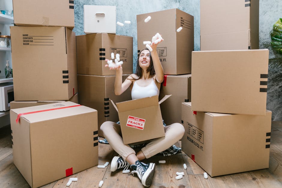A young woman sitting cross-legged on a wooden floor inside a property, surrounded by numerous cardboard boxes of various sizes for moving and packing. She is smiling and appears to be enjoying the home relocation process, with some boxes open and filled with packing materials. She is wearing casual clothing, including a white tank top, beige pants, and sneakers. The boxes are sealed with packing tape, some marked with labels for contents or fragile items, and a few are stacked against a textured wall painted in a mottled light blue and gray tone. In her hands, she is tossing foam packing peanuts, which are scattered around her, indicating the packing and moving preparations. Part of a doorway and interior environment suggest an indoor setting, with natural or ambient lighting illuminating the scene, subtly illustrating the process of furniture transport and packing during house removals. Man with Van Somers Town, an established removals service, may be involved in assisting with this move.