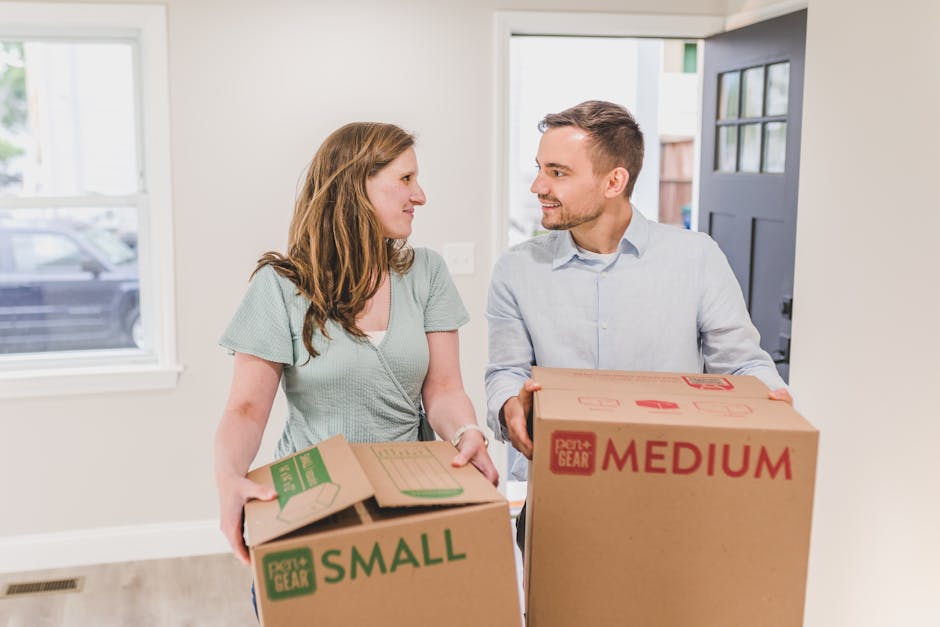 A cardboard moving box labeled with 'NAME', 'CONTENTS', 'ORDER NO.', and 'ROOM KITCHEN' written in black and blue ink. The box is sealed with clear packing tape and positioned on a paved surface outside a property, as part of a home relocation process. In the background, there are additional packed boxes and a hand truck or trolley used for transporting household items. This scene captures the loading or unloading stage of furniture transport and packing during a house removal carried out by Man with Van Somers Town, demonstrating professional moving and packing services.