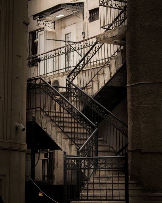 A narrow, outdoor metal staircase attached to an older building, with multiple flights connected by small landings, featuring black wrought iron railings with decorative elements. The stairs are made of concrete or stone, with some signs of weathering, and the setting includes a beige façade and a window with partially open shutters on the upper level. The staircase is situated against a brick wall on one side, and a small overhanging metal canopy is visible at the top. The image depicts an urban apartment building, where Man with Van Somers Town might carry out home relocation or furniture transport services, involving careful navigation of the confined staircase during a moving or packing and moving process.