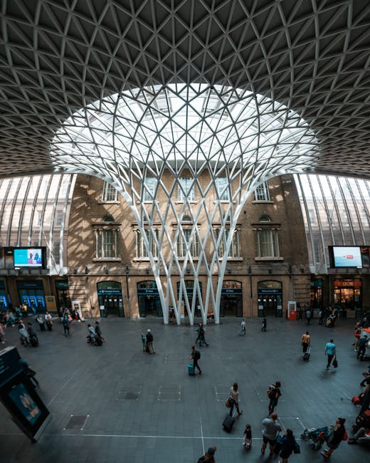 Interior view of a modern train station concourse with a high, geometrically patterned metal roof structure supported by white, tree-like columns. The spacious area is filled with numerous passengers