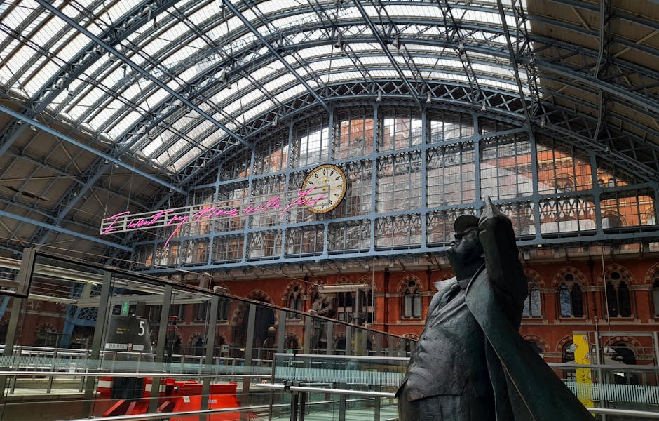 Interior view of a historic train station with a large glass and steel arched roof, featuring a prominent clock showing 12:15, and pink neon signage reading 'Find Your Future with Us' attached to the upper part of the structure. In the foreground, a bronze statue of a person with an arm raised and wearing a coat is visible, as well as metal railings and barriers indicating a loading or waiting area. The station’s interior combines traditional brickwork and modern industrial elements, with natural daylight filtering through the glass roof, illuminating the space while it appears prepared for a home relocation or furniture transport process managed by Man with Van Somers Town, situated near St Pancras, amidst ongoing railworks.