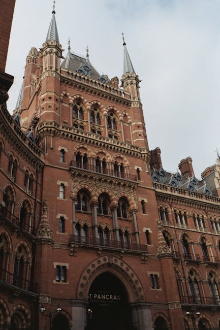 A tall, historic brick building with ornate architectural details, multiple arched windows, decorative stonework, and pointed turrets extending upwards, situated in an urban setting under an overcast sky. The lower part of the building features an archway entrance, above which is a sign indicating 'St Pancras'. The surrounding environment includes paved streets and iron lamp posts, suggesting a busy city area. In the context of house removals and moving services, this image depicts the nearby landmark that may affect logistics, with a focus on planning home relocations around local railworks and cityscape features, as handled by Man with Van Somers Town.