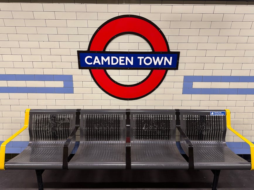 An empty seating area at Camden Town underground station, featuring a row of four metal benches with perforated seats and backs, positioned against a cream brick wall. The benches have black supporting frames with yellow armrests on each end, and a blue stripe runs along the backrests. Above the benches, a large circular London Underground sign with a red border, black outlines, and a blue bar displays the station name 'Camden Town' in white uppercase letters. The station environment is brightly lit, clean, and ready to facilitate passenger waiting during home relocation or furniture transport needs. This setting reflects the typical loading process associated with house removals and moving services, such as those provided by Man with Van Somers Town, who may utilize station access for efficient furniture transport and packing in busy city streets.