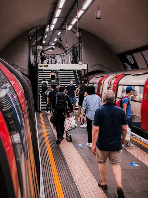 A group of passengers, some carrying backpacks and shopping bags, walking along a platform at an underground London station with curved ceilings and modern lighting. Two trains are visible on either side of the platform, with one train having its doors open. Some individuals are ascending a staircase at the end of the platform, which is also equipped with safety rails and signage. The scene depicts typical busy city transport activity, reflecting the context of urban home relocations or furniture transport logistics. This image is associated with Man with Van Somers Town, providing house removals and packing services connected to moving processes in the Somers Town to Euston Station area.
