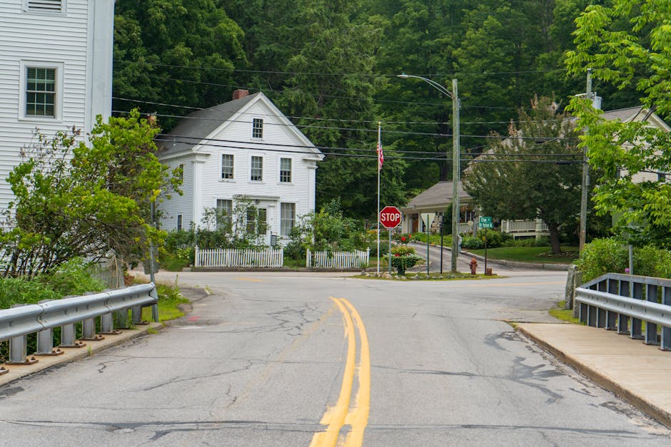 A residential street scene showing a paved road with yellow double lines and metal guardrails on both sides. In the background, there is a white, two-story house with multiple windows, surrounded by trees and greenery. The street intersects with another road at a stop sign, and additional houses and trees are visible further back. The scene appears to be in a quiet neighborhood with an overcast sky, with no vehicles or people present, capturing a typical setting for home relocation or moving logistics involving packing, loading furniture, and transportation during a house move, as handled by Man with Van Somers Town.