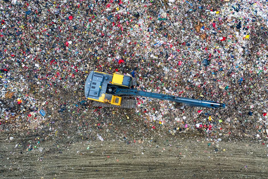 An aerial view of a small excavator with a yellow body and black arm, operating on a large area of landfill filled with mixed waste materials such as plastic bottles, paper, and other debris. The excavator is situated near the bottom center of the image, close to a dirt pathway at the edge of the waste site. The surrounding waste is densely packed, creating a textured surface of various colours and materials. This scene illustrates the process of waste disposal often encountered during house removals and furniture transport, highlighting the importance of proper waste management and compliance with waste disposal laws in NW1. Man with Van Somers Town occasionally manages waste removal as part of their house relocation services, ensuring efficient and legal disposal of unwanted items during moving projects.