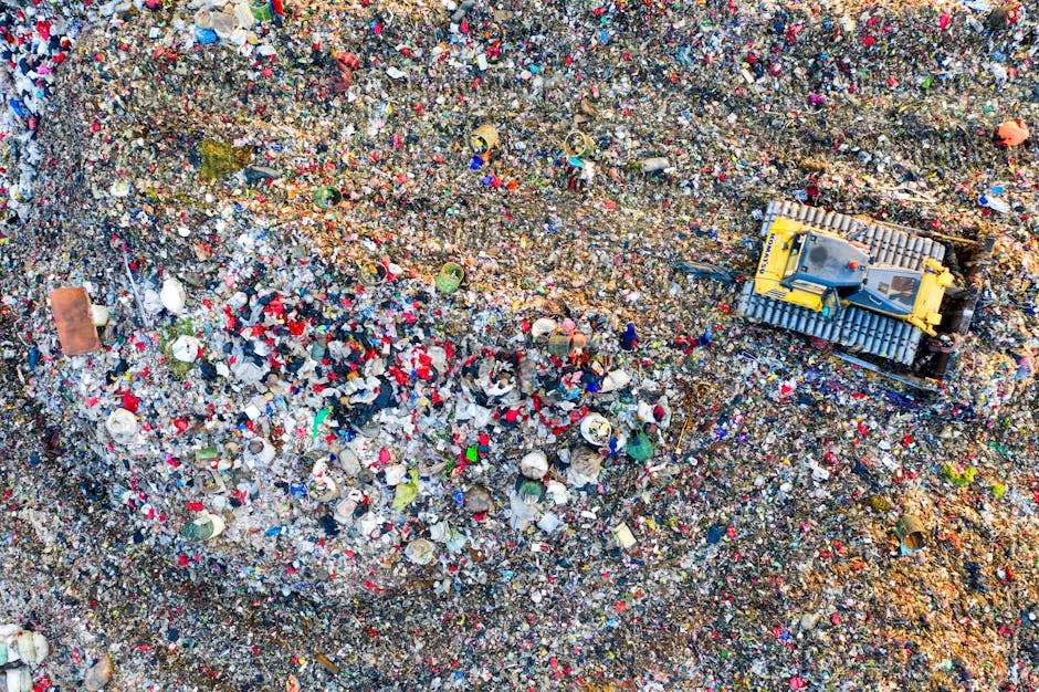 An aerial view of a small excavator with a yellow body and black arm, operating on a large area of landfill filled with mixed waste materials such as plastic bottles, paper, and other debris. The excavator is situated near the bottom center of the image, close to a dirt pathway at the edge of the waste site. The surrounding waste is densely packed, creating a textured surface of various colours and materials. This scene illustrates the process of waste disposal often encountered during house removals and furniture transport, highlighting the importance of proper waste management and compliance with waste disposal laws in NW1. Man with Van Somers Town occasionally manages waste removal as part of their house relocation services, ensuring efficient and legal disposal of unwanted items during moving projects.